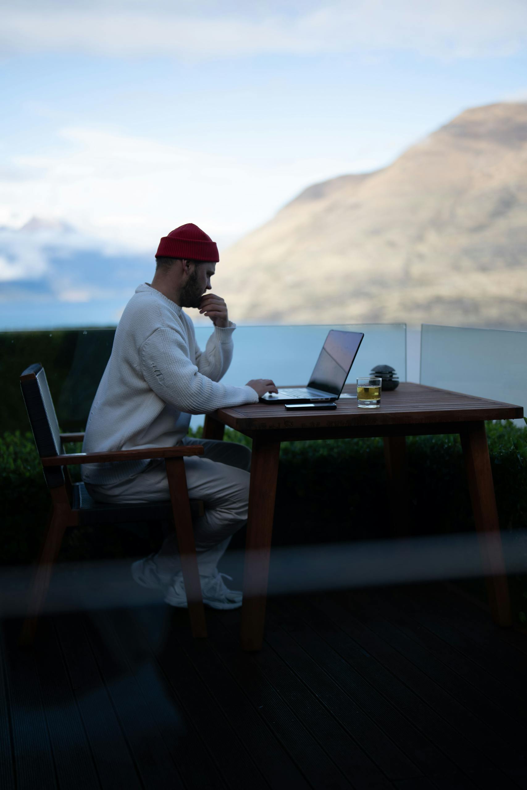 A man in casual attire works on a laptop on a terrace with stunning mountain views in Queenstown, New Zealand.
